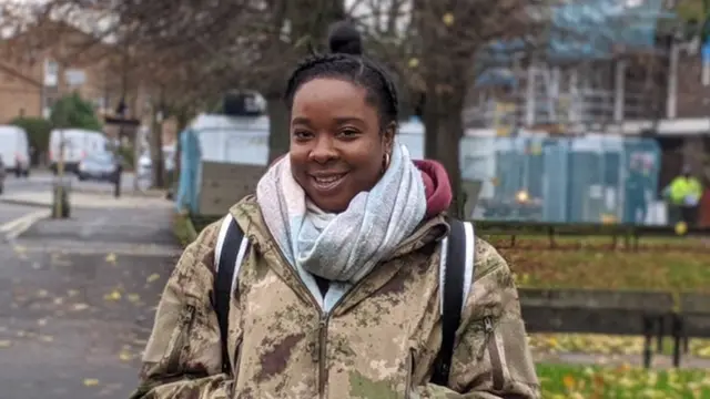 Sharell est debout, souriant à la caméra, les mains dans ses poches