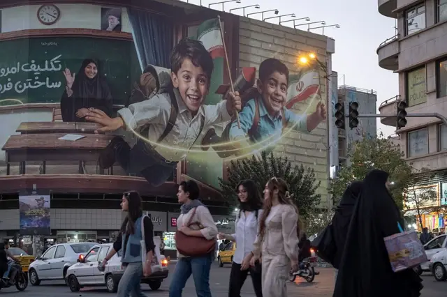 یک بیلبورد بزرگ در تهران که دو دانش‌آموز پسر را نشان می‌دهد که خندان هستند در حالت پرواز هستند، یک معلم محجبه و نیمکت‌ها پشت سر این دو دانش‌آوز است. روی بیلبورد نوشته شده: تکلیف امروز ساختن فردا. در گوشه بیلبورد تصویر موشک نیز دیده می‌شود. عابران زن بدون حجاب و با حجاب در حال عبور از مقابل این بیلبورد هستند