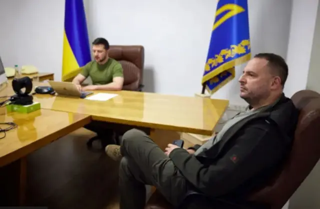 На передньому плані Єрмак сидить у кріслі і дивиться у бік, на задньому плані - Зеленський сидить за світло коричневим столом 