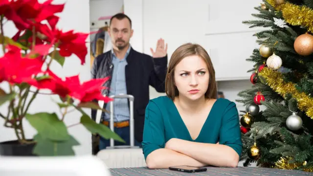 Kobieta siedzi przy stole. Obok niej widać ustrojoną choinkę i poinsecję. Mężczyzna podnosi rękę w geście pożegnania i wychodzi z domu.