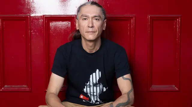 ဒေါ်အောင်ဆန်းစုကြည်ရဲ့ သားအငယ် ကင်မ်အဲရစ် (ခ) ကိုထိန်လင်း။ မြန်မာနိုင်ငံမြေပုံ၊ထောင်နံရံနဲ့ မိခင်ဖြစ်သူပုံကို ဒီဇိုင်းဆွဲထားတဲ့ တီရှပ်အနက်ကို ဝတ်ဆင်ထားပါတယ်။