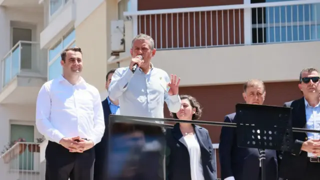 Özgür Özel otobüs üzerinde konuşuyor. Yanında eşi ve takım elbiseli kişiler var.