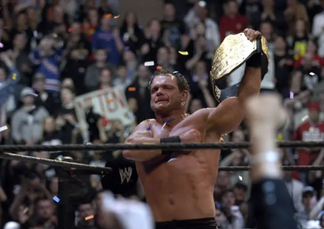 Chris Benoit, ex luchador de la WWE, sostiene visiblemente emocionado, el título de campeón mundial de lucha libre en medio del ring. 