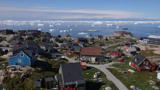 Um conjunto de casas, algumas pintadas de vermelho e azul, cercadas por grama e ligadas por estradas asfaltadas, em frente ao mar pontilhado de icebergs na Baía de Disko, 15 de julho de 2024 em Ilulissat, Groenlândia