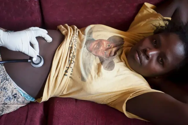 Une femme enceinte couché dans un canapé, habille en t-shirt jaune, se fait consulter par un médécin dont on voit la main gantée.