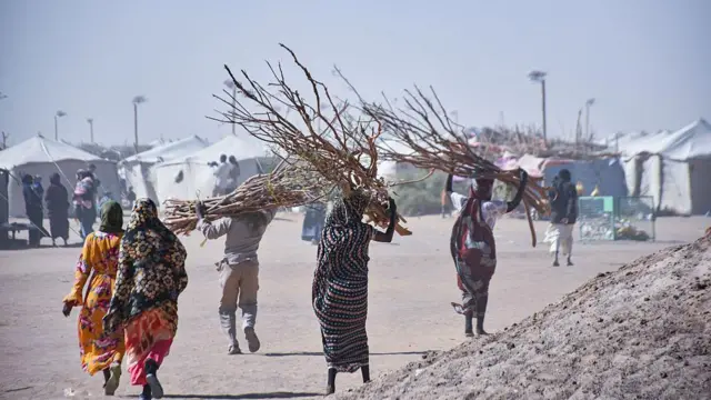 تظهر نساء سودانيات من الخلف باللباس التقليدي يحملن حطباً، وعدد آخر من الرجال يحمل أحدهم حطباً. كل شخص في الصورة يظهر ماشياً لوحده قرب عدد من الخيام. الصورة التقطت نهاراً.