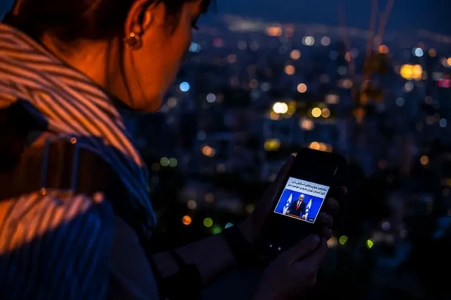 زنی در حال نگاه کردن به موبایل خود است، نمایی از شب شهر در پسزمینه دیده می‌شود