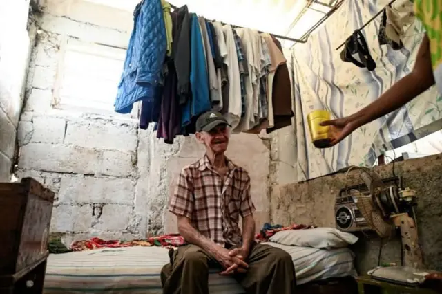 Senhor pardo idoso em uma cama simples em um cômodo de paredes não rebocadas, com roupas penduradas em um varal acima dele