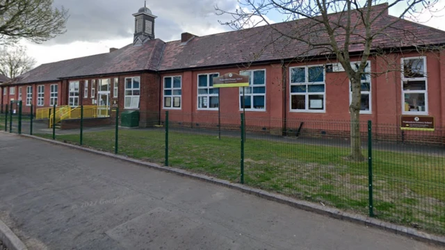 The single storey red-brick school building sits behind a thin metal fence with grass leading up to the school building.