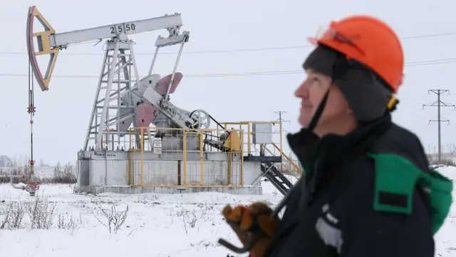 Зимний пейзаж. На заднем плане нефтяной станок-качалка. На переднем справа человек в профиль в рабочей одежде и оранжевой каске.