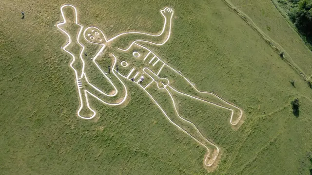 Una toma aérea de la silueta blanca de un hombre desnudo con un garrote, dibujada en una colina de hierba verde. Se ven personas dispersas alrededor de la figura; algunas están agachadas en el suelo y otras caminan mientras llevan herramientas en las manos.