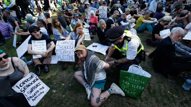 Полицейский задерживает сидящего мужчину на акции 6 сентября среди десятков мирно восседающих на траве протестующих с одинаковыми плакатами "Я против геноцида, я поддерживаю Palestine Action"