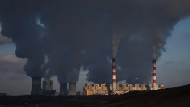 Dym i para unoszą się z czerwono-białych kominów i chłodni kominowych. Promienie słońca odbijają się od kominów i budynków.