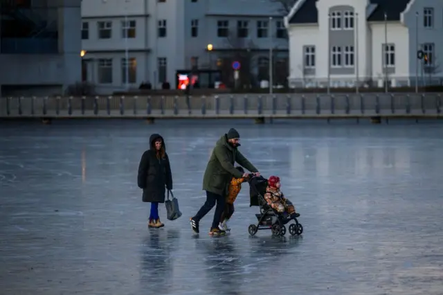 Família patinando no gelo, com casas em bom estado ao fundo