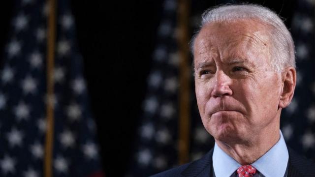 Joe Biden looks into the distance with US flags behind him.