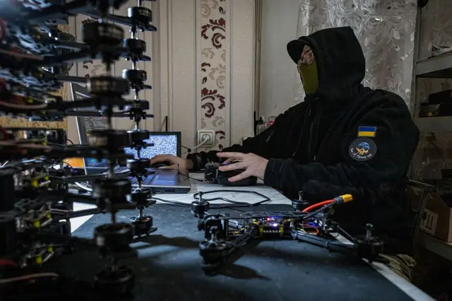 За ноутбуком сидить людина одягнена у темний худі з нашивкою з українським прапором, а також у маску, що закриває нижню частину обличчя. На передньому плані видно стос квадрокоптерів та окремі деталі безпілотників, розкладені на столі. На задньому плані — стелажі з коробками та обладнанням, а також вікно з напівпрозорою фіранкою.