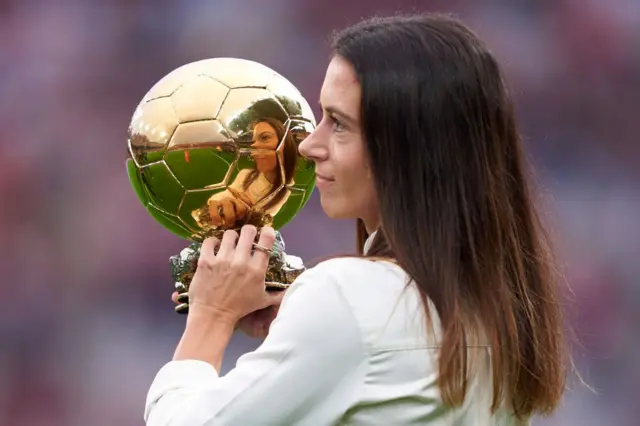 Женщина с распущенными волосами держит в руках золотой мяч, в котором отражается ее лицо