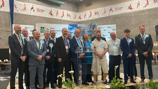 Un grupo de hombres, principalmente con trajes oscuros, y un par de mujeres, la mayoría con escarapelas azules de Reform UK, sonriendo en Kettering Leisure Village.