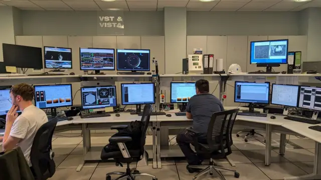 Dos personas observan pantallas de computador en lo que parece ser una sala de control.