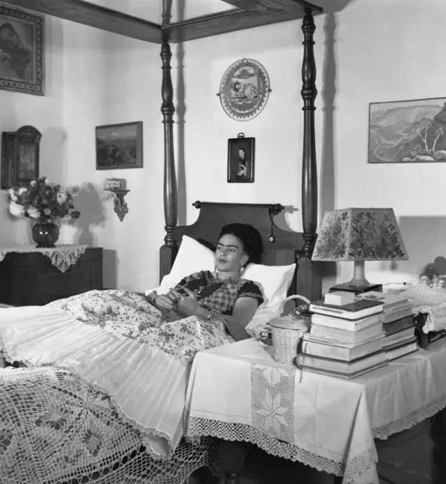 Frida Kahlo, con aretes, el cabello recogido y su ropa tradicional, está acostada en su cama. Su cabeza y espalda están sobre una almohada blanca. Al lado tiene una mesa con una lampara, una cesta y varios libros. Al otro lado, hay un florero con flores y en la pared hay cuadros.