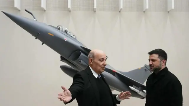 クリーム色の壁に掲げられた灰色の戦闘機の写真を背に、黒いスーツ姿の男性が両手を動かしながら話すのを、黒い服装のゼレンスキー氏が神妙な表情で聞いている