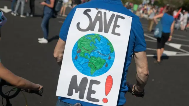 Homem com um cartaz com um desenho da Terra escrito "save me" (me salve,site analise esportivainglês)