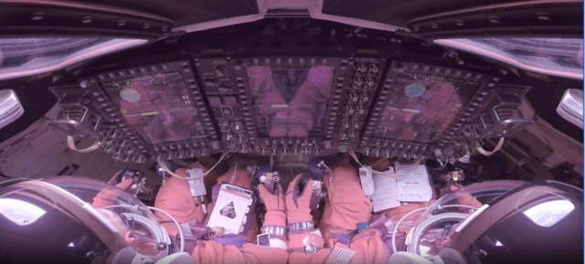 这张影像从两名阿提米丝二号太空人的肩后视角向前拍摄，他们身穿橘色加压太空衣，被牢牢固定在猎户座乘员舱的座椅上。两人戴着透明头盔罩，面向一整面高耸、发光的显示萤幕，以及密集排列的开关与旋钮。戴着手套的双手放在操控杆与检查清单旁。画面边缘可见弧形窗户、缆线与设备，使整个舱室显得紧凑而高度技术化。