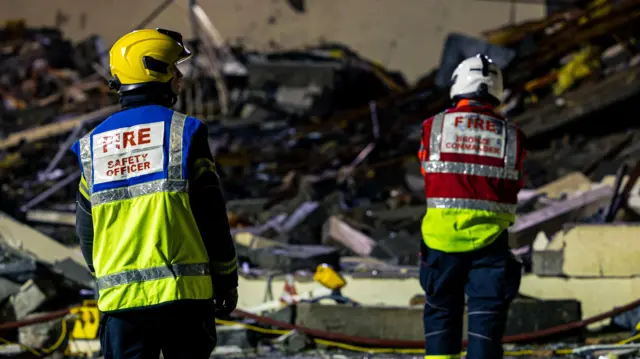 Dos bomberos con chalecos y cascos de alta visibilidad se encuentran frente a una gran pila de escombros de una estructura derrumbada. En uno de los chalecos se lee "OFICIAL DE SEGURIDAD CONTRA INCENDIOS". La escena está tenuemente iluminada, lo que sugiere que es de noche o hay poca luz.