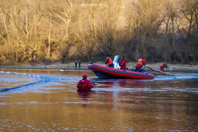 Спасатели в защитных костюмах работают в неглубокой речной воде рядом с моторной лодкой. Один из них стоит по пояс в воде. В реке видна длинная плавучая заграждающая лента, которую команда разворачивает для сдерживания загрязнения.