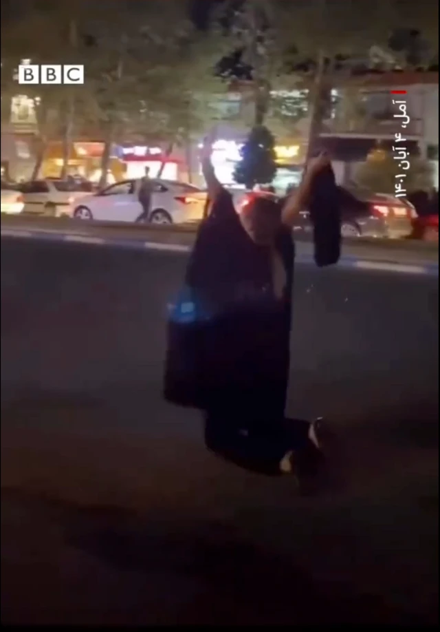 تصویر زن معترض در آمل که روی آسفالت نشسته است، روسری خود در در آورده و دستانش را بالا برده است