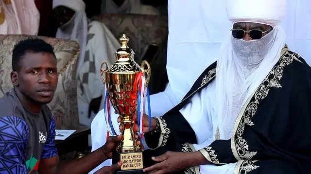 A Dambe fighter from the House of Arewa accepts the Emir Ado Bayero Dambe Championship trophy from His Highness the Emir Aminu Ado Bayero
