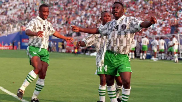 Une image de la Coupe du monde 1994 aux États-Unis montre trois joueurs nigérians célébrant un but. Daniel Amokachi, auteur de l’action, y danse avec exubérance, les bras écartés, dans un mouvement rythmé des hanches, tandis qu’il claque des doigts et ajuste son équilibre après avoir inscrit un but décisif.