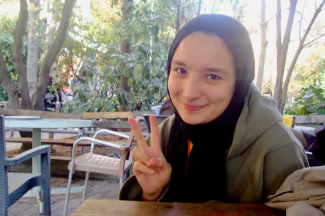 Zehra cafe gibi bir açık ortamda masada oturmuş sağ eliyle V işareti yapmış, kameraya gülümsüyor. 