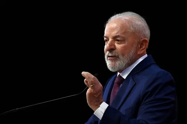 Lula é um homem branco de cabelos grisalhos. Ele está falando ao microfone. Ele usa um terno azul