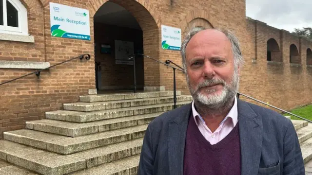 El líder del Consejo del Condado de Devon, Julian Brazil, viste una camisa rosa claro y un suéter burdeos de cuello en V bajo una chaqueta azul marino. Está de pie al pie de las escaleras que conducen a la entrada principal del Ayuntamiento.