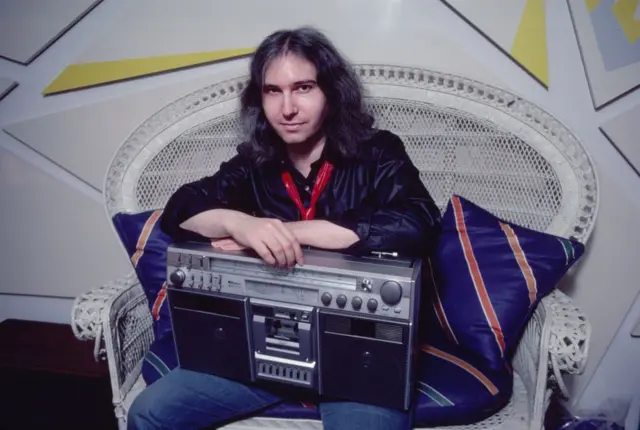 Jim Steinman, un hombre de entre 30 y 40 años con el cabello largo, una camisa negra y una casetera en las piernas, está sentado y mira a la cámara.