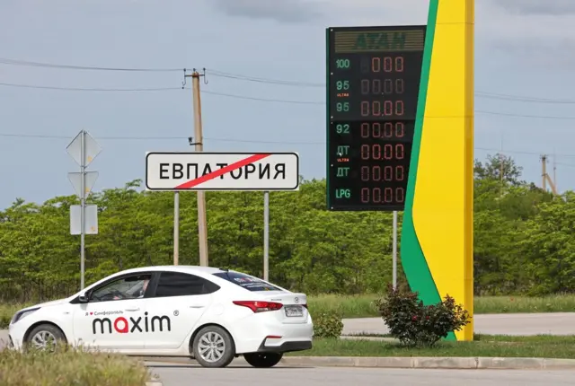 На заднем фоне темно-синее небо, зеленые кусты и столбы с праводами. На среднем плане знак выезда из Евпатории. Рядом стоит погасшее табло бензозаправки "Атан". На переднем плане стоит белая легковая машина сервиса такси "Максим".
