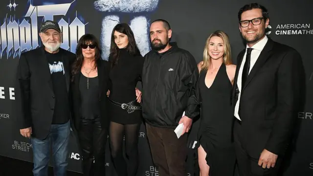 黒字に白と灰色で「Spinal Tap II」と書かれたボードの前に、全員が黒い服装で並んだ家族。