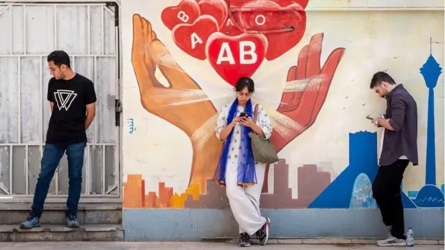 Tres personas están de pie frente a un colorido mural pintado en una pared. El mural muestra dos manos grandes, una roja y otra naranja, sosteniendo figuras con los grupos sanguíneos "A, B, AB y O" escritos en su interior.