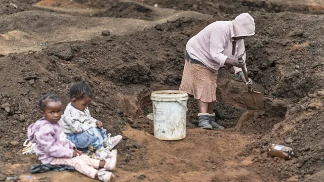 Mwanamke anatumia sepeto kujaza ndoo na udongo karibu na watoto wake huku wachimbaji wengine wakichimba mashimo wakitafuta dhahabu katika kipande cha ardhi nje ya Springs.