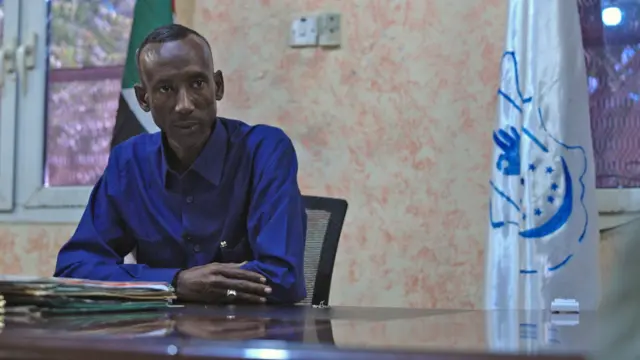 Tijani Karshoum nọ ọdụ n'oche, yi uwe elu na-acha anụnụ anụnụ. Enwere ọkọlọtọ ọcha na anụnụ anụnụ n'aka nri.