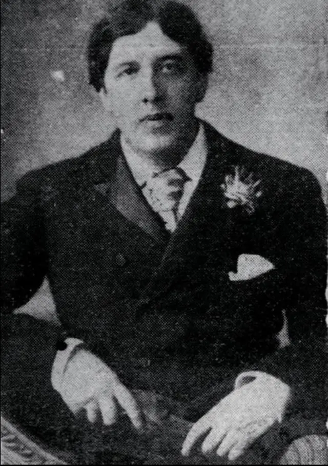 En una fotografía en blanco y negro, el famoso poeta, autor y dramaturgo Oscar Wilde mira ligeramente a la derecha de la cámara. Lleva un traje extravagante con un pañuelo asomando del bolsillo superior de la chaqueta, con lo que parece ser un broche prendido justo encima.