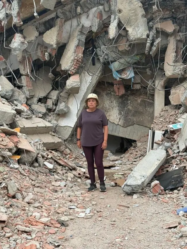 María Lourdes Pérez, vestida con un pantalón y camisa marrones y un gorro beige. Detrás de ella hay una montaña de escombros, hormigón, piedras, partes de una edificación