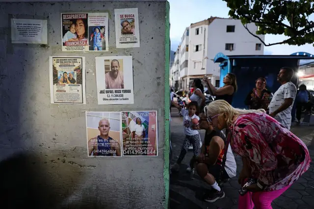 Varios carteles con fotos de personas desaparecidas están pegados a una pared y varias personas se agachan a mirar la información que incluyen. 