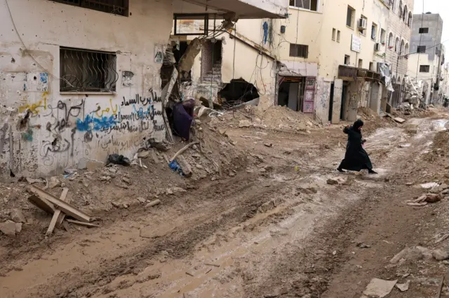 امرأة فلسطينية تعبر طريقاً بعد تجريفه بجوار المباني المتضررة، خلال عملية عسكرية إسرائيلية في مخيم جنين للاجئين في الضفة الغربية المحتلة، في 3 مارس/آذار 2025