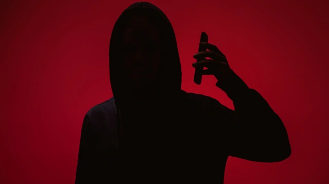 ظل شخص يرتدي قبعة رأس ويمسك هاتفاً محمولاً قرب أذنه، بخلفية حمراء موحّدة، مع إخفاء كامل لملامح الوجه