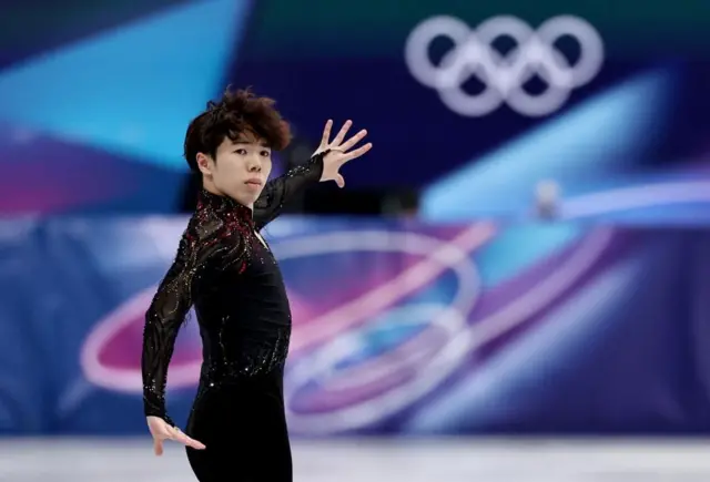 黒い衣装の佐藤がリンクに立ち、右横を見ている。左手は伸ばして指を開き、右手は腰の位置に下げている。背景に五輪マークなどがぼんやり見える