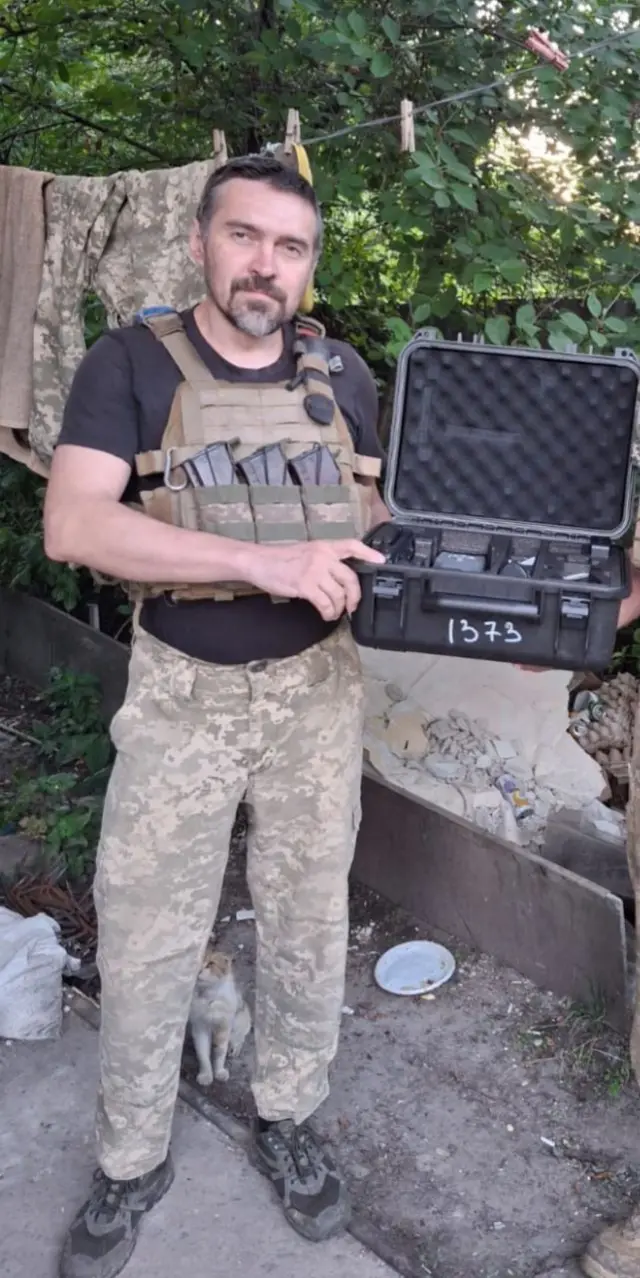 У повний зріст стоїть Сергій Капуста, батько Дмитра, у камуфляжних військових брюках, у броніку, чорній футболці. Сергій тримає військову валізу обладнанням. Вона відкрита