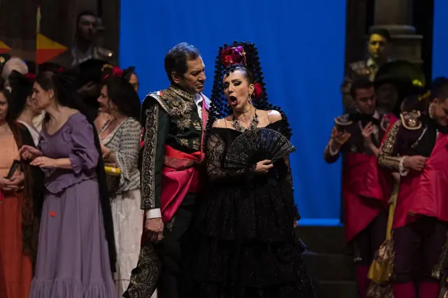 Sahnede opera söyleyen bir kadın ve erkek. Arkada figüranlar. Herkes dönem kostümleri giymiş. 