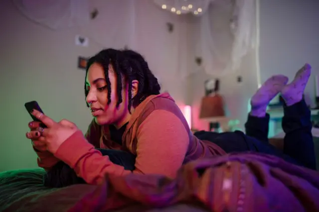 Una estudiante en la cama boca abajo con un teléfono celular en la mano. 